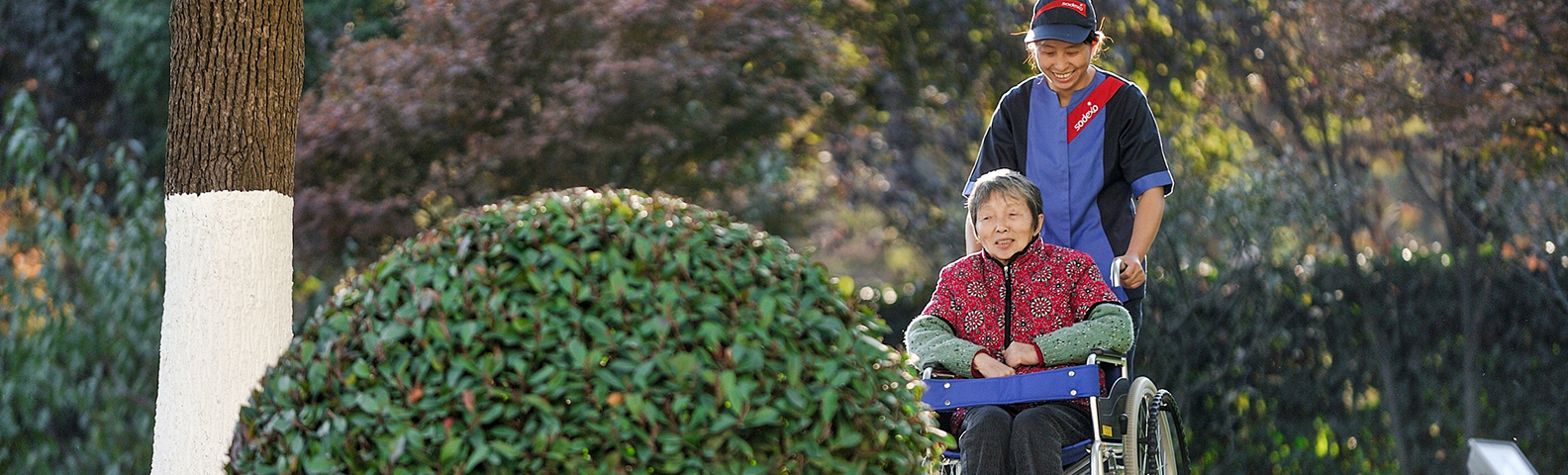 Sodexo employee pushing an elderly woman in a wheelchair through a garden
